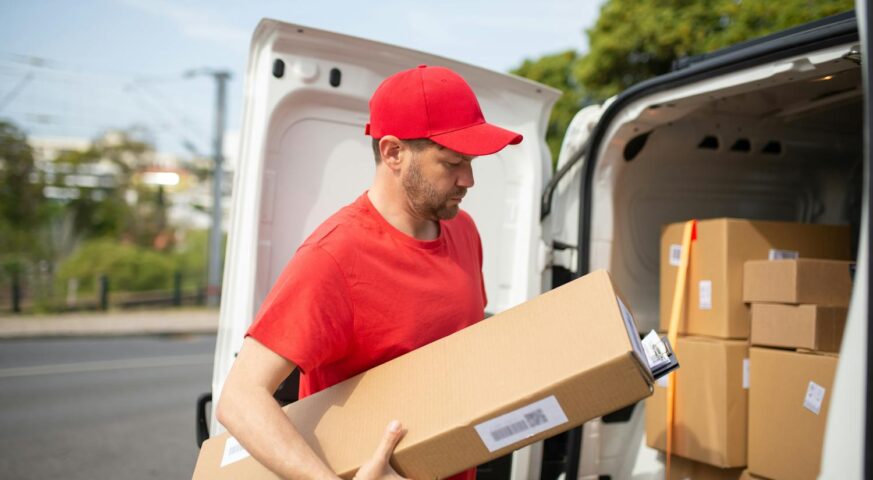 Mann in roter Uniform trägt einen Karton aus einem mit Paketen beladenen Lieferwagen