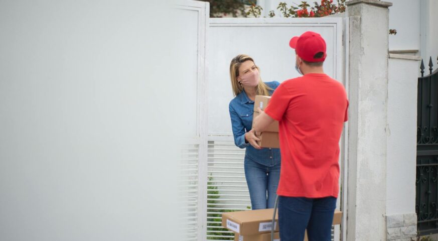 Eine Frau mit Maske nimmt ein Paket von einem Lieferfahrer in rotem T-Shirt und Kappe an ihrer Haustür entgegen