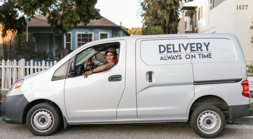 Ein Lieferfahrer sitzt in einem weißen Lieferwagen mit der Aufschrift "Delivery Always On Time", geparkt vor einem Wohnhaus